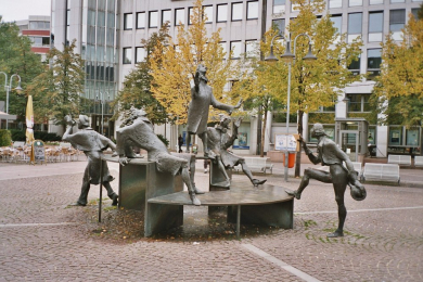 Brunnen aus Bronze auf einem gepflasterten Platz mit Darstellung einer aus fünf Personen bestehenden Figurengruppe.