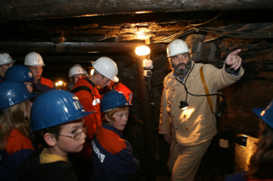 Ein Bergmann mit Schutzhelm führt eine Gruppe Kinder durch das Bergwerk.
