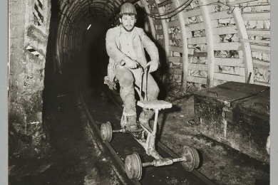 Fotografie eines Bergmanns an einer Streckenabzweigung unter Tage auf einem schienengebundenen Grubenrad fahrend.