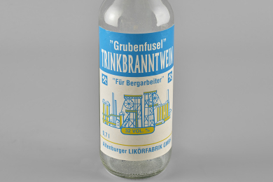 Leere Flasche aus klarem Glas mit einem blau-weißen Etikett. Der Titel lautet „Grubenfusel“; Trinkbranntwein „Für Bergarbeiter“. Auf dem Etikett ist eine abstrakte Darstellung eines Bergwerks mit zwei Fördergerüsten sowie Aufbereitung und weiteren Tagesanlagen zu sehen. Hersteller ist die Altenburger Likörfabrik GmbH.