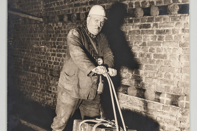 Bergmann unter Tage mit einem einspurigen Schienenrad auf Gleisen stehend. Auf dem Schienenrad sind zwei Kästen. Das Schienenrad hat die Form eines Tretrollers.