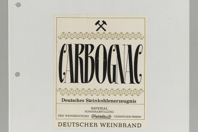 Zwei Etiketten, aufgeklebt auf einem Blatt Papier. Die Bezeichnung auf dem Etikett lautet „Carbognac“; Deutsches Steinkohlenerzeugnis. Die Etiketten sind mit einer goldfarbenen Linie umrahmt sowie mit Schlägel-und-Eisen-Symbolen dekoriert. Hersteller ist die Weinbrennerei Dujardin & Co., Uerdingen/Rhein.