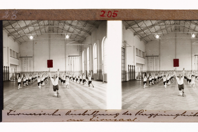 Bergjungleute, halb kniend in einer Turnhalle, wiederholen die ihnen durch den Lehrer gezeigten gymnastischen Übungen.