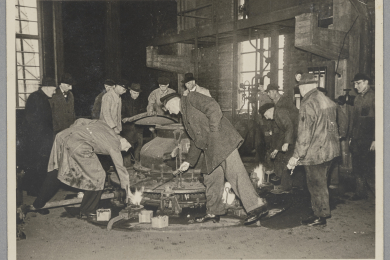 Vierzehn Personen in einer Industriehalle um einen Schwelkessel versammelt, von denen zwei, darunter Theodor Haber, den Kessel zünden.