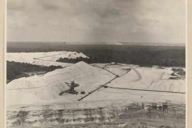 Luftbild eines Tagebaus; im Zentrum ein Bagger in weißer Mondlandschaft; dahinter Regenwald. 