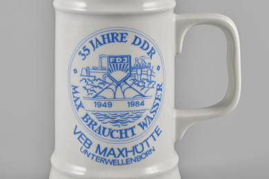 Porzellanhumpen, weiß mit blauen, umlaufenden Linien oben und unten, mittig in Blauen Linien eine Vignette, darin stilisierte Darstellung der Maxhütte und der Wasserleitung sowie das Logo der FDJ, dazu die Beschriftung „35 Jahre DDR, 1949 1984, Max braucht Wasser“ und unter der Vignette „VEB Maxhütte Unterwellenborn“.