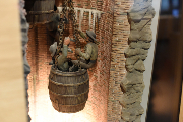 Detailansicht des Dioramas Seilfahrt mit Förderkübeln, Anfertigung der Modellbauwerkstatt des damaligen Bergbau-Museums Bochum, 1941. Kübel mit drei Bergleuten in Arbeitskleidung, alle drei sind mit brennenden Froschlampen ausgestattet. Ein Bergmann mit erhobenem Arm im Kübel stehend, die anderen beiden auf dem Rand des Kübels sitzend und stehend.