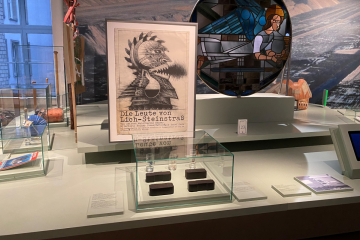 Blick in einen Ausstellungsraum mit Vitrinen, Plakaten und Ausstellungsobjekten