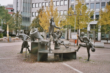 Brunnen aus Bronze auf einem gepflasterten Platz mit Darstellung einer aus fünf Personen bestehenden Figurengruppe.
