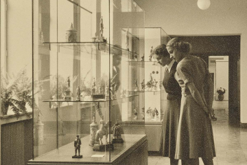 Sepia-Foto, das zwei Frauen vor einer Vitrine mit Kunstgegenständen zeigt.