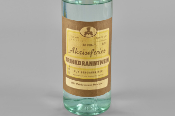 0,7-Liter-Flasche aus klarem Glas mit einer klaren Flüssigkeit und goldfarbenem Schraubverschluss. Auf dem braun-gelben Etikett ist der Inhalt als Akzisefreier Trinkbranntwein für Bergarbeiter beschrieben. Weiterverkauf wird strafrechtlich verfolgt. VEB Weinbrennerei Meerane.