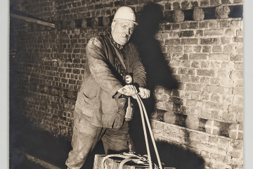 Bergmann unter Tage mit einem einspurigen Schienenrad auf Gleisen stehend. Auf dem Schienenrad sind zwei Kästen. Das Schienenrad hat die Form eines Tretrollers.