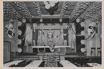 Großer Raum in einem Lokal mit einer Bühne und Tischen und Stühlen davor. Girlanden, Lampions und Masken sind an der Decke und an der Bühne angebracht.