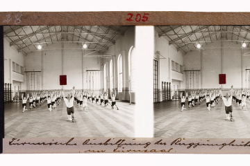 Bergjungleute, halb kniend in einer Turnhalle, wiederholen die ihnen durch den Lehrer gezeigten gymnastischen Übungen.