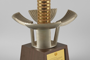 Zu sehen ist ein Objekt aus Metall auf einem Holzsockel. Der Holzsockel trägt vorne eine Metallplakette mit einem Text in englischer Sprache. Der Metallaufsatz erinnert entfernt an eine silberfarbene Blüte, die sich um einen goldfarbenen Zylinder öffnet.