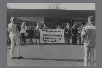 Eine Gruppe von älteren und jüngeren Frauen, teils mit Kindern, demonstriert vor einer Zufahrt, in der Mitte ein Transparent mit der Aufschrift „Wir kämpfen für die deutsche Steinkohle. Fraueninitiative Sophia-Jacoba“. Sie werden von Journalisten gefilmt. 