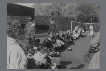 Eine Gruppe von Frauen und Kindern sitzt aufgereiht auf dem Asphalt, dazwischen drei stehende Polizisten. 