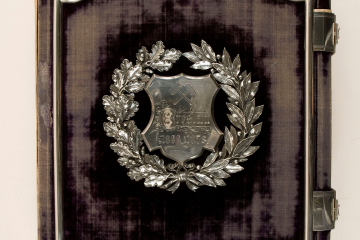 Das Bild zeigt den Deckel des samtbezogenen Fotoalbums mit aufwändigem Silberschmuck sowie zwei silbernen Schlössern auf der rechten Seite. Innerhalb eines silbernen Blätterkranzes befindet sich ein Emblem, auf dem unter dem Signet mit Schlägel und Eisen der Name „Breslau“ und die Jahreszahlen „1866“ und „1878“ eingraviert sind