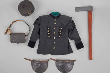 Eine schwarze Uniformjacke mit Messingknöpfen umgeben von einem Filzhut, einem Beil, einem Paar Knieschützern und einer Tasche