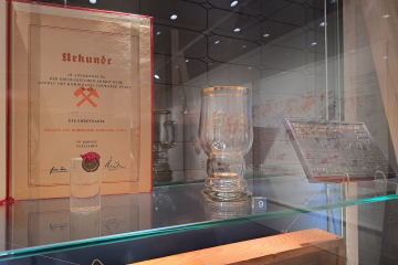Ein Trinkglas mit Henkel und Aufschrift in einer Ausstellungsvitrine neben einer Urkunde, einer Ehrennadel und einer Plakette