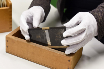 Ein Museumsmitarbeiter mit Baumwollhandschuhen hebt vorsichtig Glasplatten-Aufnahmen aus einem Holzkasten