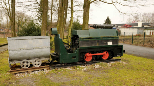 Eine Grubenlok mit Grubenwagen steht auf einem Rasen vor Bäumen.
