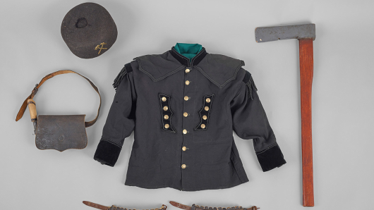 Eine schwarze Uniformjacke mit Messingknöpfen umgeben von einem Filzhut, einem Beil, einem Paar Knieschützern und einer Tasche