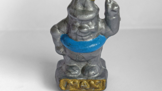Kunststofffigur eines silbrigen Männchens mit spitzem Hut auf einem Sockel mit erhobenem linkem Arm, Flammen auf dem Kopf und einem blauen Schwimmreifen um den Bauch. Auf dem Sockel steht gold hervorgehoben MAX.