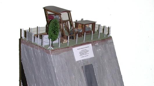 Prismatisch, einseitig abgeschrägtes Modell aus Sperrholz, farbig gefasst, mit Einsichten von drei Seiten in Schacht, Querschlag und Flözstrecke; auf der Oberseite Darstellung der übertägigen Anlagen; Kennzeichnung der Schichten durch Außenbemalung; vorne Schild mit Beschriftung.